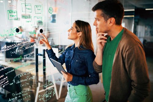 Eine Frau bespricht mit einem Mann aufgeschriebene Notizen auf einem Glas-Whiteboard.
