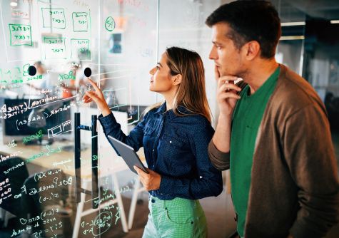 Eine Frau bespricht mit einem Mann aufgeschriebene Notizen auf einem Glas-Whiteboard.